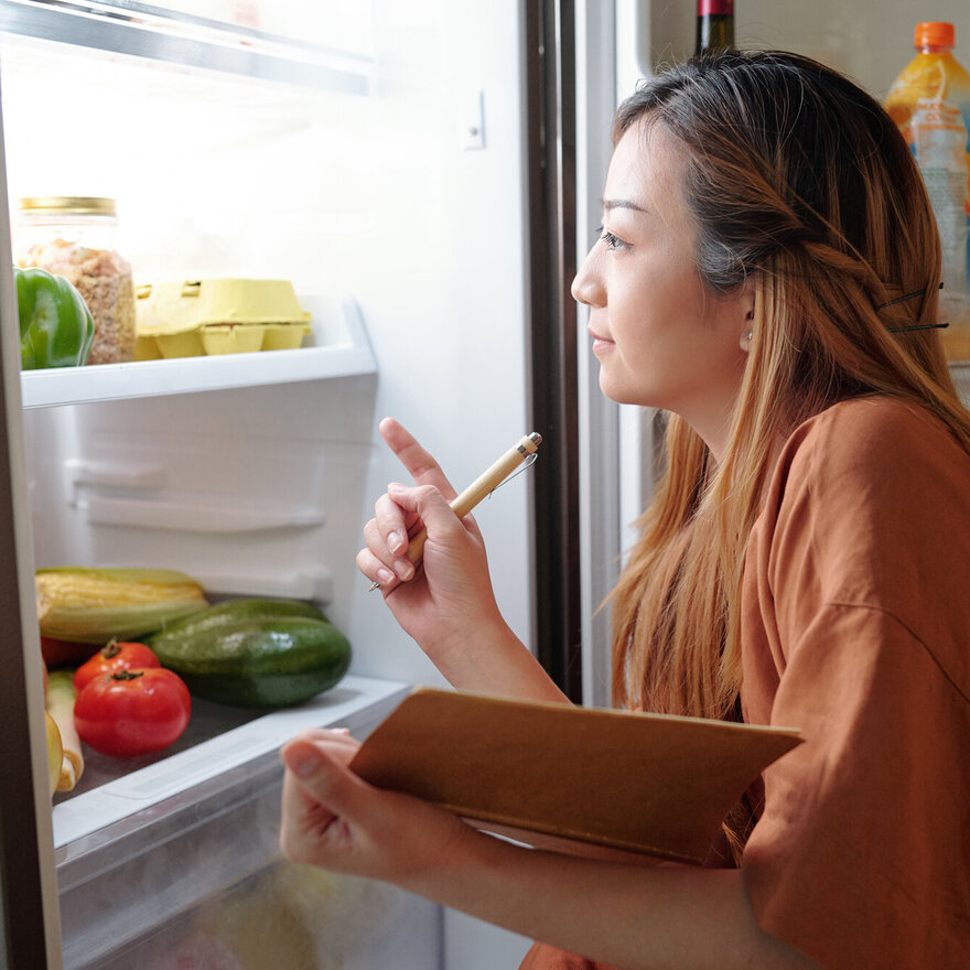 Pensive woman with notepad checking groceries in her fridge and writing down a list of products she need to buy
