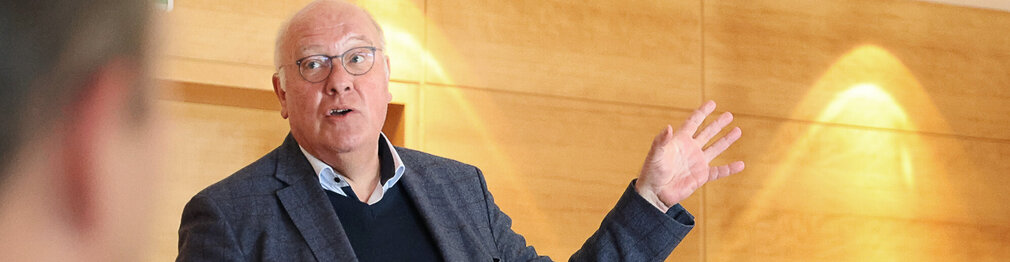 Portraitfoto von Wolfgang Rigger; er spricht und deutet mit einer offenen Hand nach rechts