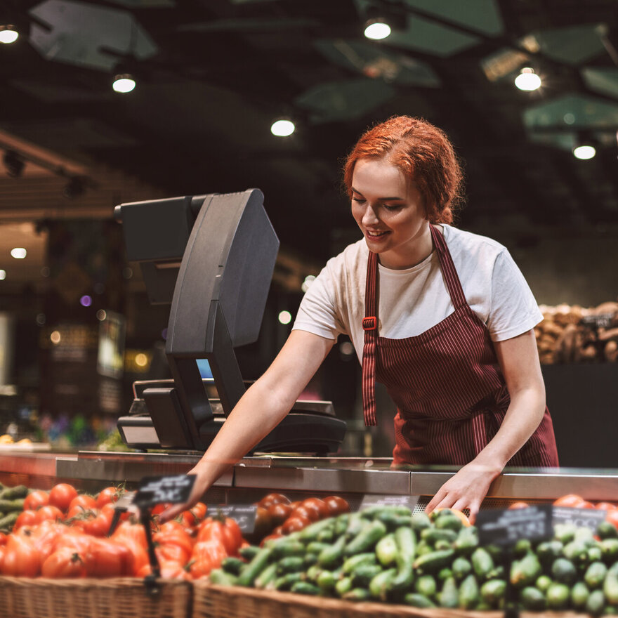 Young smiling seller in apron behind counter with vegetables hap